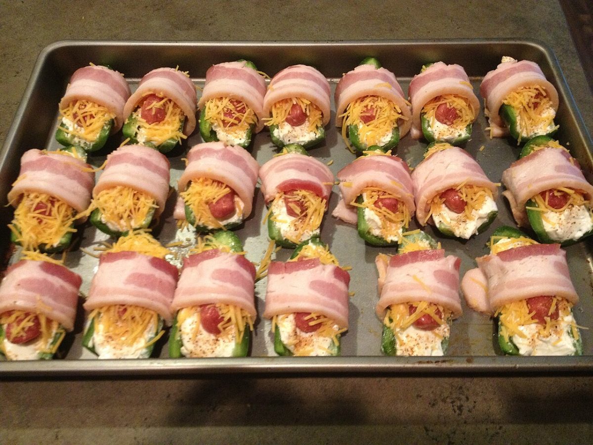 A tray of jalapeno halves stuffed with cream cheese and cheddar, wrapped in bacon, ready to bake