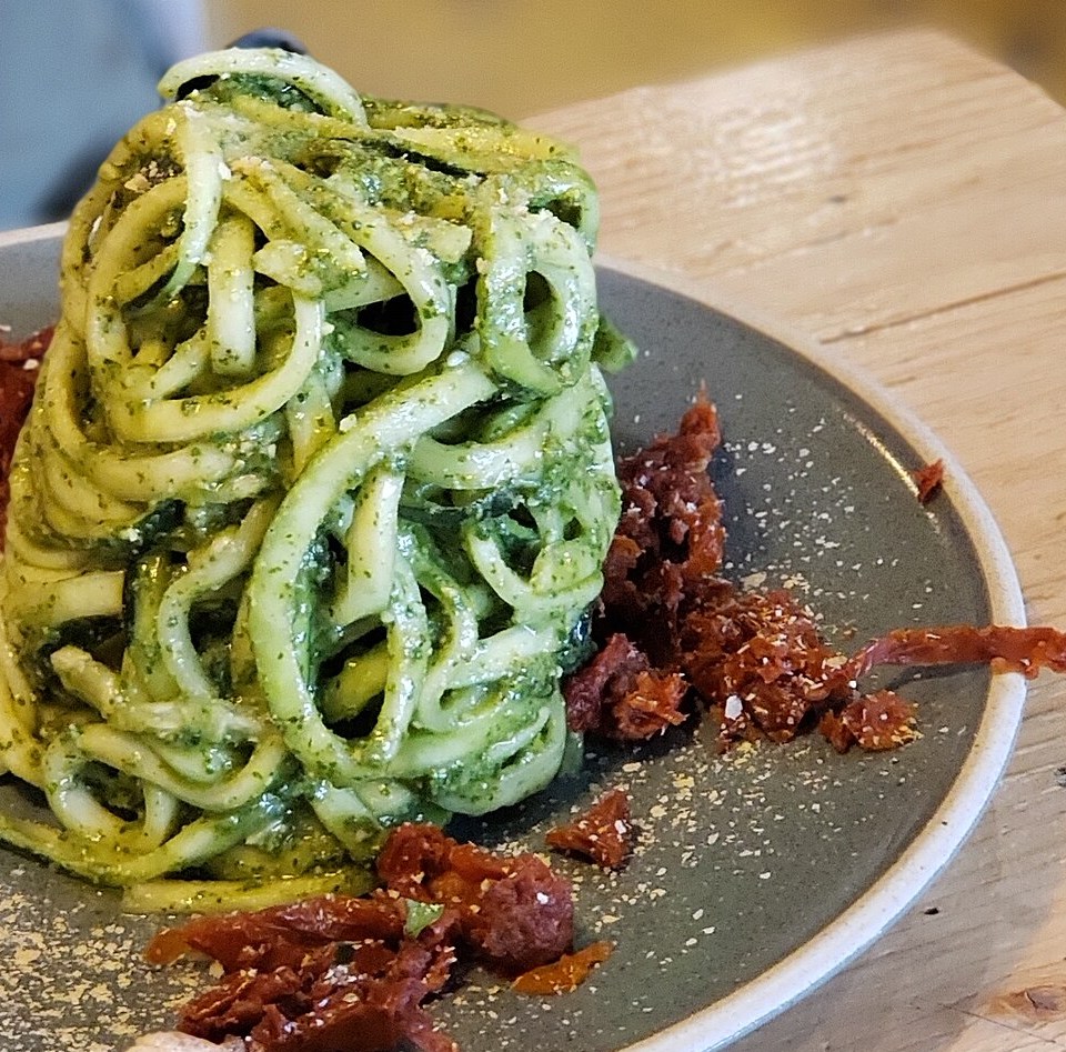 A tall nest of zucchini noodles coated in green pesto with sun-dried tomatoes on a gray plate