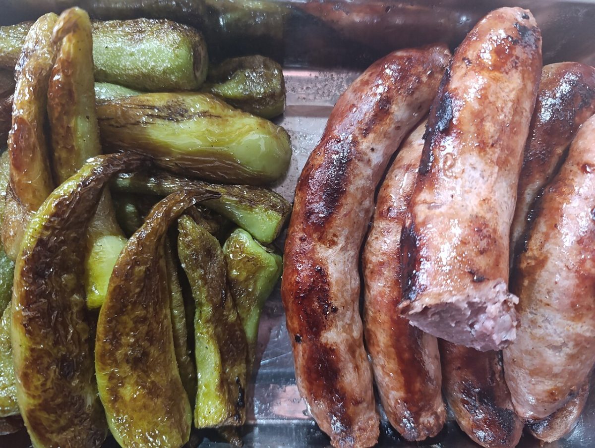 Browned sausages next to roasted green peppers on a metal baking tray