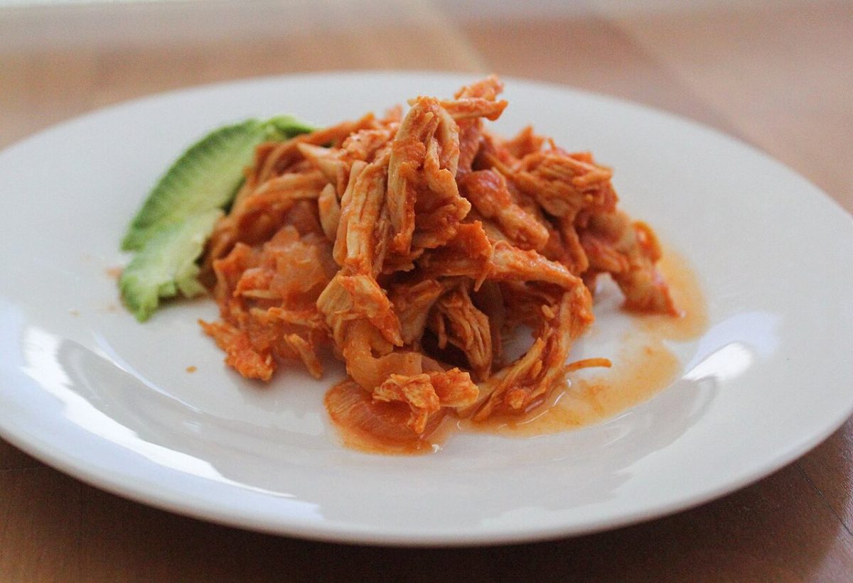 Shredded chicken in a red tomato sauce on a white plate with avocado slices